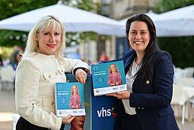 Nicole Boissier steht links, rechts steht Dr. Carolin Lehberger, beide halten ein Programmheft in der Hand und lächeln in die Kamera