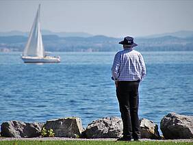 Mann steht am Ufer und schaut auf Bodensee, im Hintergrund Berge, Segelboot auf dem See