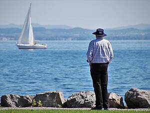Mann steht am Ufer und schaut auf Bodensee, im Hintergrund Berge, Segelboot auf dem See