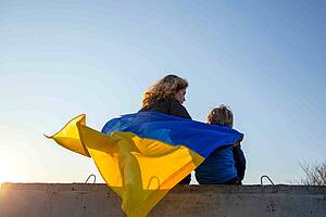 Eine Frau und ein Kind sitzen auf einer Mauer und tragen die ukrainische Flagge als Mantel auf dem Rücken