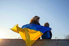 Eine Frau und ein Kind sitzen auf einer Mauer und tragen die ukrainische Flagge als Mantel auf dem Rücken