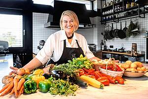 Die Köchin Léa Linster steht in der Küche, vor ihr liegt Gemüse und Obst auf dem Tisch