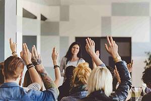 Man sieht mehrere Menschen von hinten in einer Klasse, die den Arm heben, um sich zu melden. Vorne steht eine Lehrerin